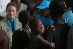 On 22 March 2019 in Beira in Mozambique, (centre front) a young boy looks toward the camera as (background left) UNICEF Executive Director Henrietta H. Fore speaks with internally displaced people as she visits a secondary school used to shelter evacuees from Cyclone Idai. Tropical cyclone Idai, carrying heavy rains and winds of up to 170km/h (106mp/h) made landfall at the port of Beira, Mozambique’s fourth largest city, on Thursday 14 March 2019, leaving the 500,000 residents without power and communications lines down. On 23 March 2019, according to initial government estimates, 1.8 million people across the country, including 900,000 children, have been affected by the cyclone which slammed into the country last week. However, many areas are still not accessible and UNICEF and partners on the ground know that the final numbers will be much higher.  UNICEF is concerned that flooding, combined with overcrowded conditions in shelters, poor hygiene, stagnant water and infected water sources, is putting them at risk of diseases like cholera, malaria and diarrhoea. Initial assessments in Beira indicate that more than 2,600 classrooms have been destroyed and 39 health centers impacted. At least 11,000 houses have been totally destroyed. In Beira, Fore visited a school which had turned into a shelter for displaced families. Classrooms were converted into overcrowded bedrooms with limited access to water and sanitation. A UNICEF warehouse was severely damaged in the cyclone, causing the loss of essential supplies that had been pre-positioned before the cyclone made landfall. Cyclone Idai started as a tropical depression in Malawi, where it forced families from their homes into churches, schools and public buildings. Nearly half a million children are affected. After Mozambique, the cyclone moved to Zimbabwe where it caused significant damage to schools and water systems.  Children need to regain access to health, education, water and sanitation, and to heal from the